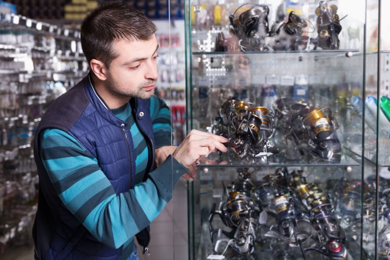 Young man choosing a fishing reel for rod in the sports shop